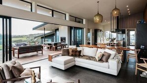 A modern living room with a sectional sofa, a dining area, and large windows offering a view of the outdoors. - Tradewinds Retreat by Waiheke Pure - Views views views, pool, spa, luxurious spacious living (Waiheke Island)