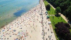 Une plage bondée où les gens se prélassent au soleil et se baignent, un ciel dégagé et, au loin, une colline boisée.