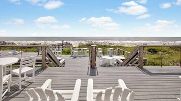 A wooden deck with white chairs and tables, leading to a beach.