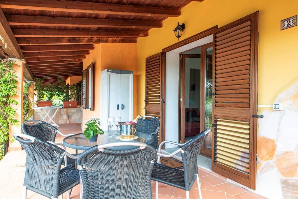 Une terrasse couverte, meublée de meubles en osier, avec une table sur laquelle se trouvent une bouteille et des verres, et une porte dotée de volets.