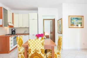 A modern kitchen with white cabinets, a coffee maker, and a dining area with patterned chairs and a wooden table. - Résidence Villa Smeralda (Telti)