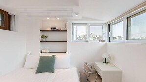 Quarto moderno com cama, escrivaninha e cadeira, janela com vista para a cidade e estante com objetos decorativos.