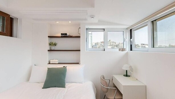 Une chambre moderne avec un lit, un bureau et une chaise, une fenêtre donnant sur la ville et une étagère ornée d’objets décoratifs.