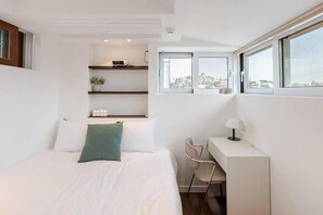 Un dormitorio moderno con cama, escritorio y silla, una ventana con vista a la ciudad y un estante con objetos decorativos.