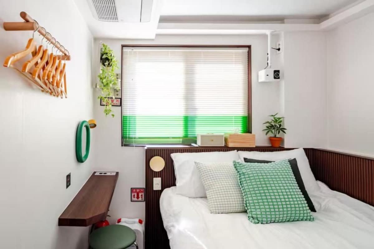 Una camera da letto con un letto, un comodino, una mensola in legno a parete, una finestra con persiane, una pianta in vaso e una telecamera fissata al muro.