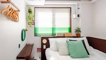 A bedroom with a bed, a bedside table, a hanging wooden rack, a window with blinds, a potted plant, and a wall-mounted camera.