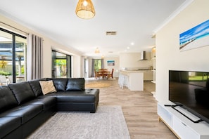 A modern living room with a black leather sofa, a flat-screen TV, and a dining area with wooden chairs and a table. - Sandpiper House (Broadwater)