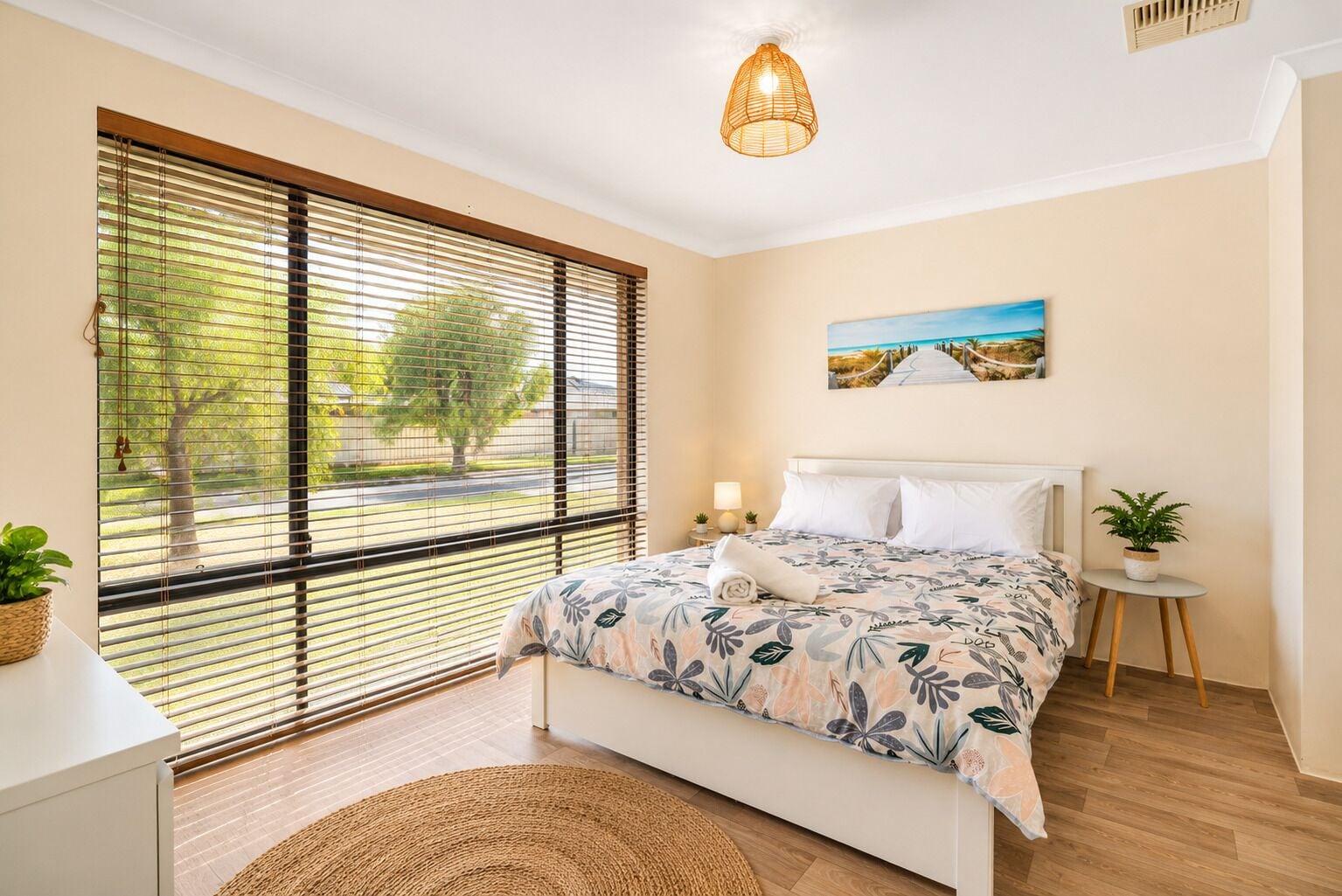 A bedroom with a bed, a window with blinds, a bedside table with a lamp, a small round table with a plant, and a painting on the wall.