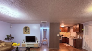 Una sala de estar moderna con un televisor de pantalla plana, un sofá y una mesa de centro. Un pasillo conduce a una cocina con armarios de madera y una encimera.