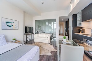 Ein modernes Hotelzimmer mit einem Bett, einer Sitzecke, einem Glastisch, einer Küchenzeile und einem großen Fenster.