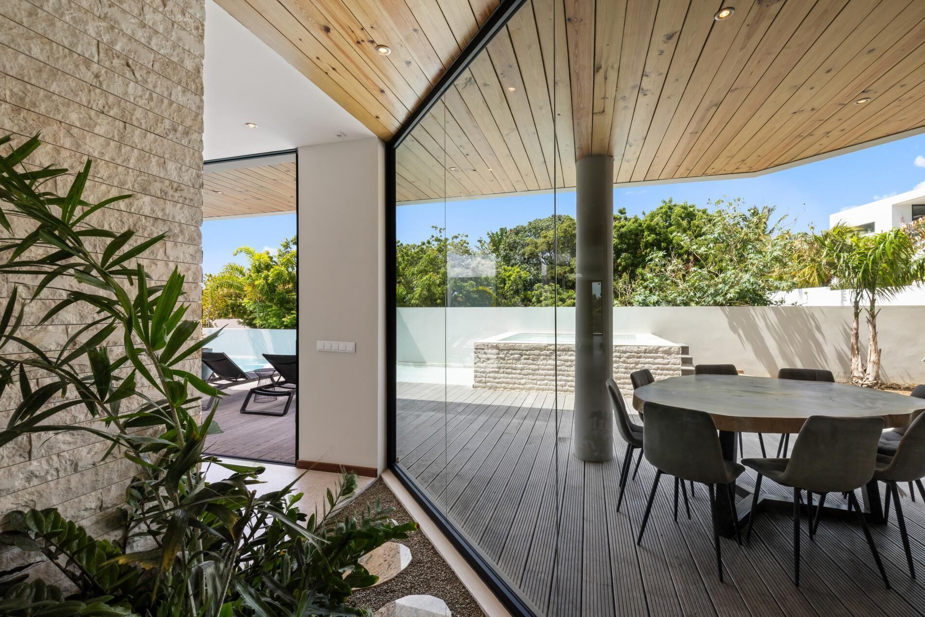 Een moderne eethoek buiten, met een glazen wand, houten plafond en uitzicht op een zwembad en groen.