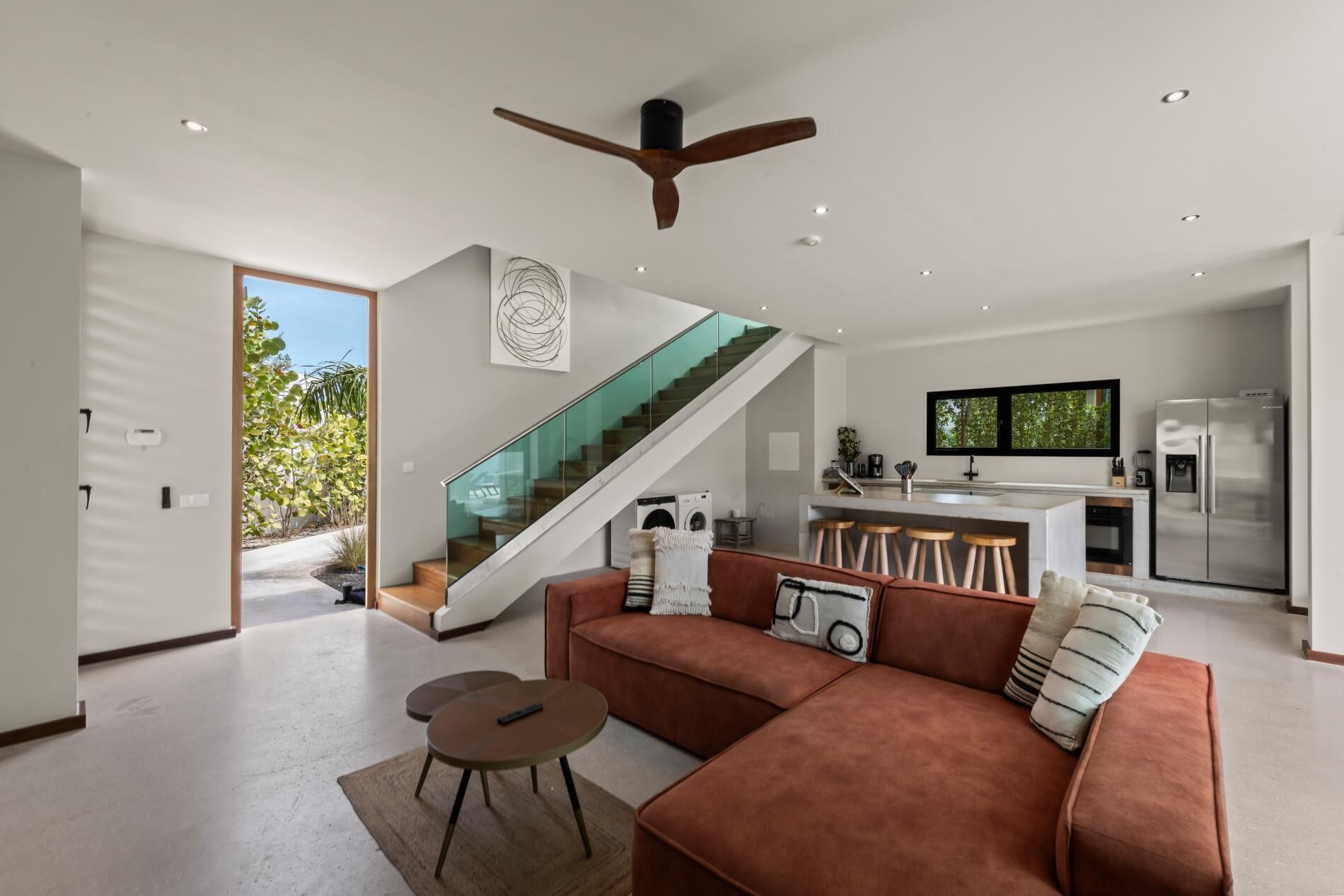 A modern living space with a sofa, a coffee table, a kitchen area with a refrigerator, and a staircase leading to an outdoor area.