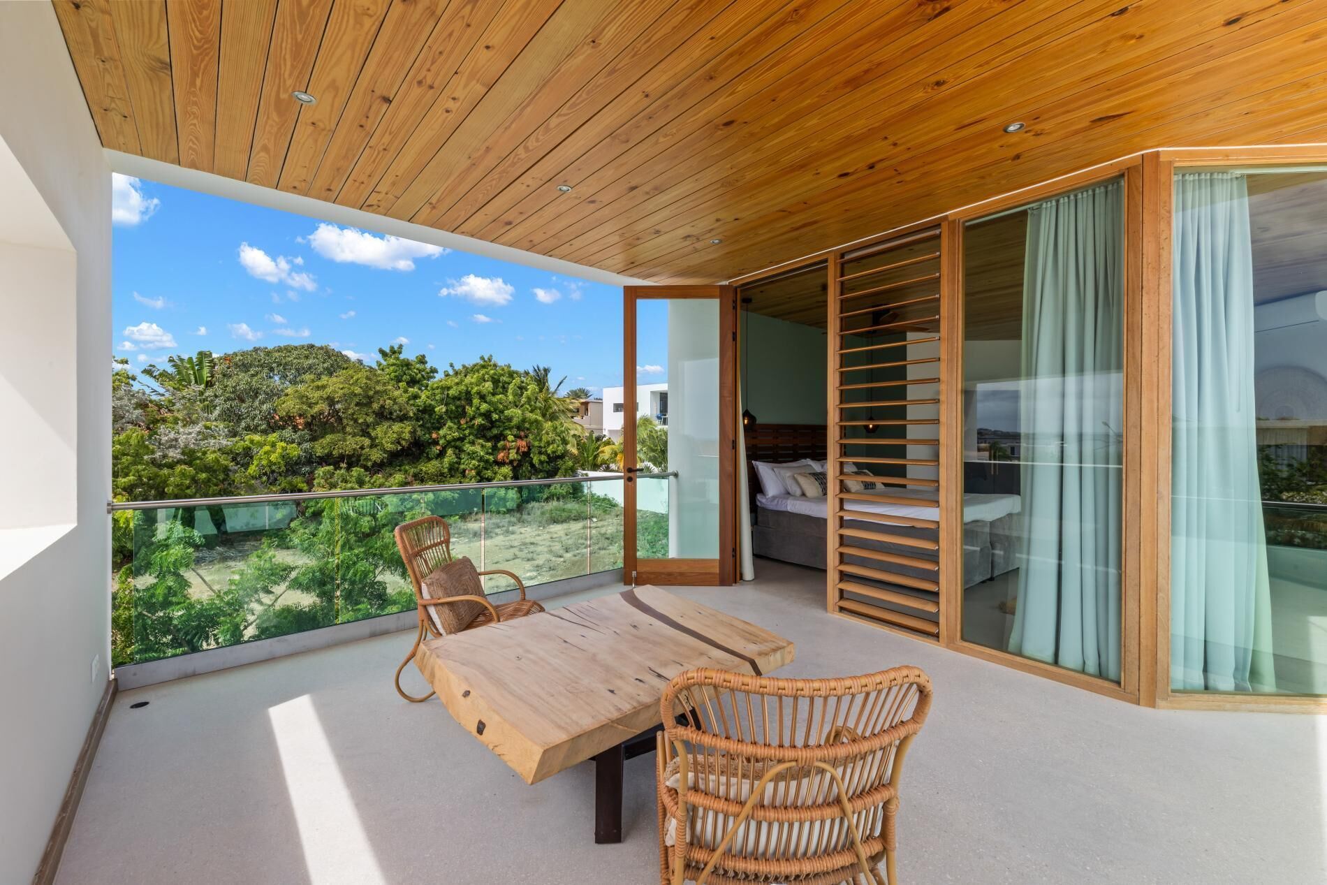 Un balcon avec une table et des chaises en bois, un lit avec une tête de lit en bois et une vue sur la verdure.