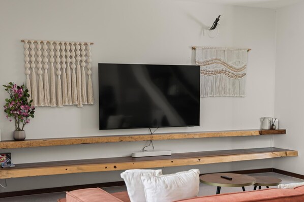 A modern living room with a flat-screen TV mounted on a wooden shelf, decorative macramé wall hangings, a potted plant, and a coffee table.