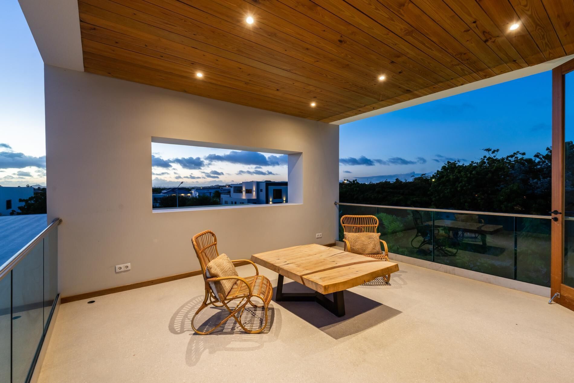 A balcony with a wooden ceiling, a table, two wicker chairs, and a view of the cityscape at dusk.