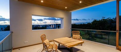A balcony with a wooden ceiling, a table, two wicker chairs, and a view of the cityscape at dusk.