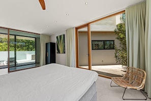 Une chambre avec un grand lit, une porte-fenêtre coulissante donnant sur un balcon et un fauteuil en osier.
