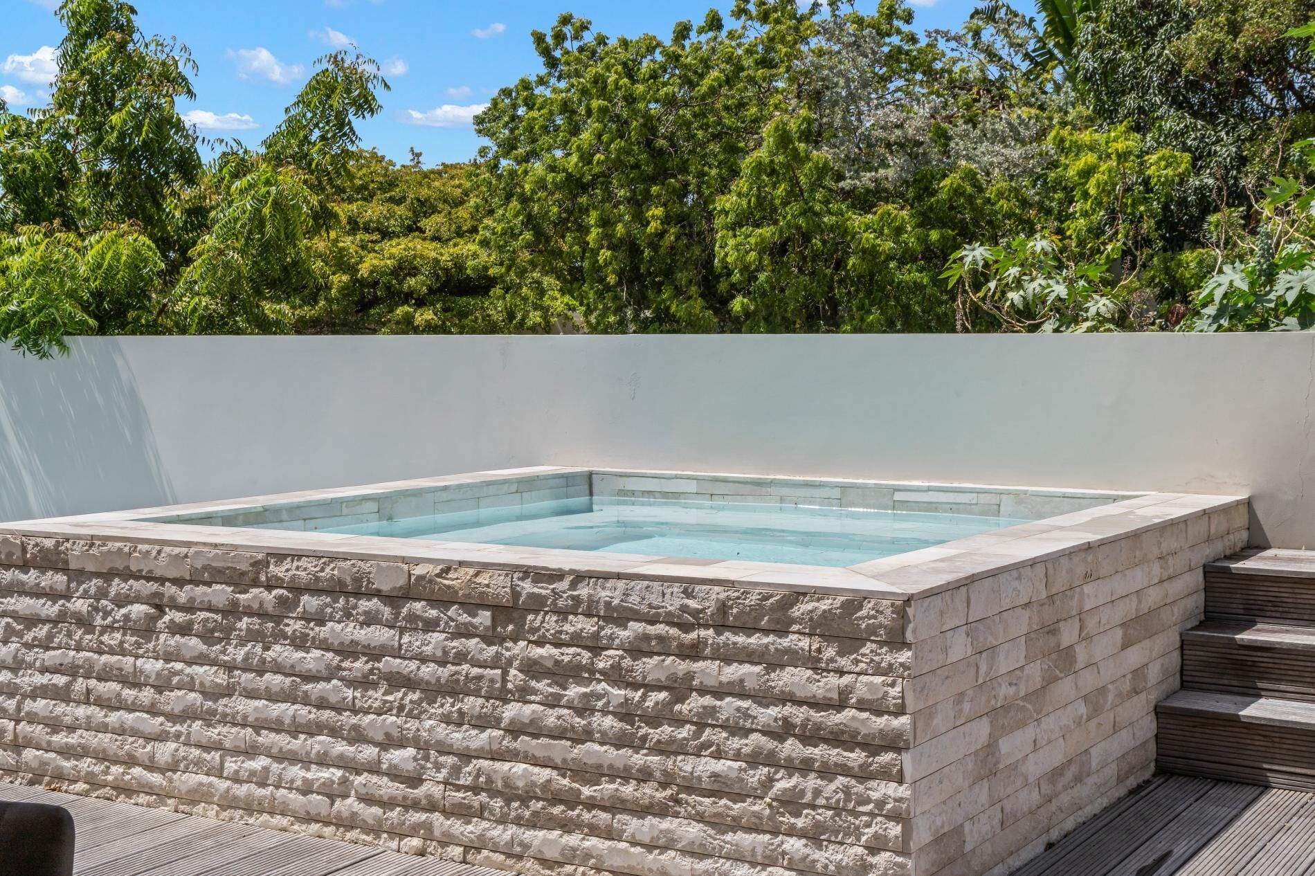 A rectangular hot tub with a stone exterior, surrounded by a white wall and a wooden deck.