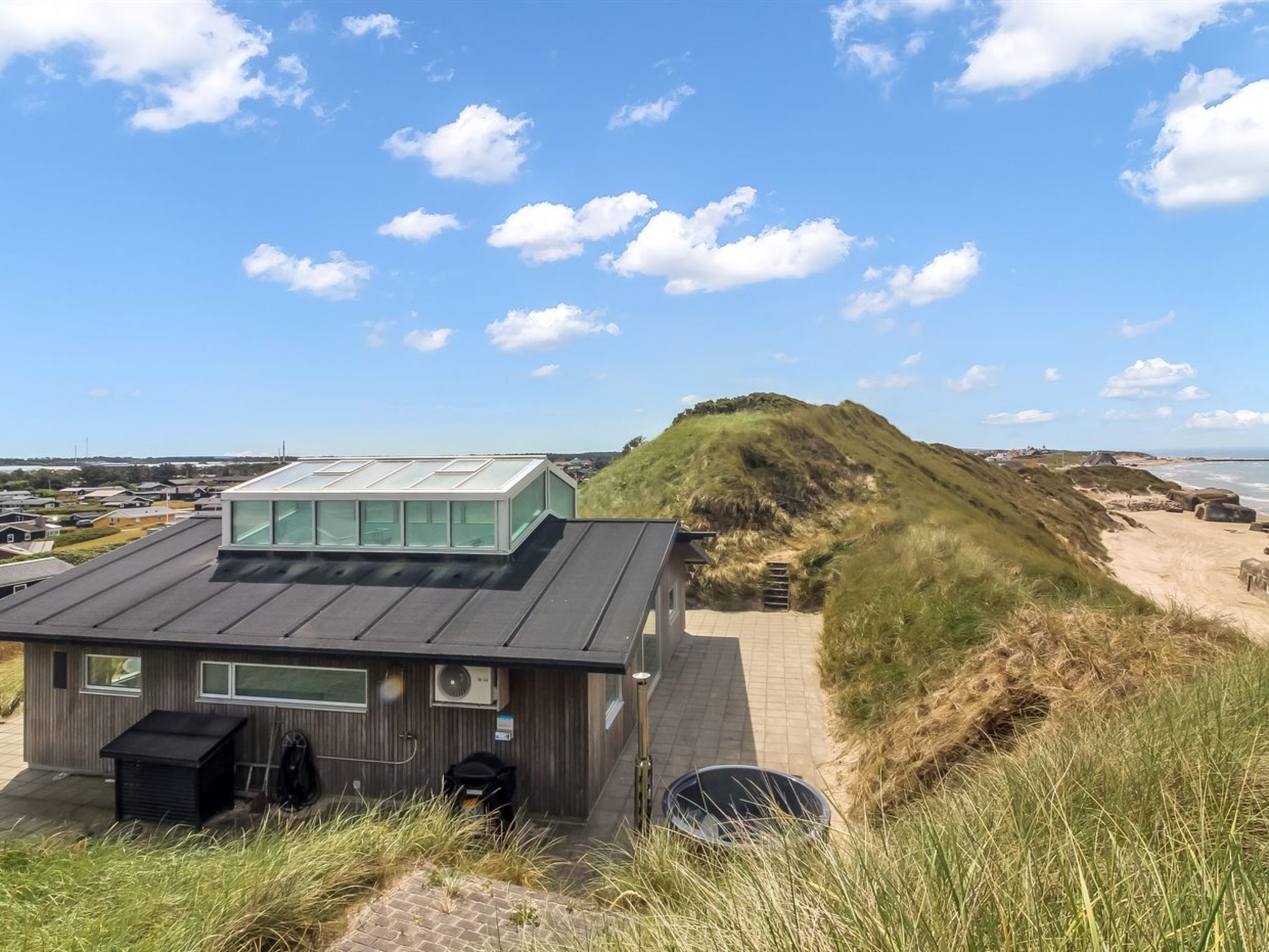 Une maison moderne avec un toit plat et de grandes fenêtres, située sur une colline verdoyante qui offre une vue sur la plage.