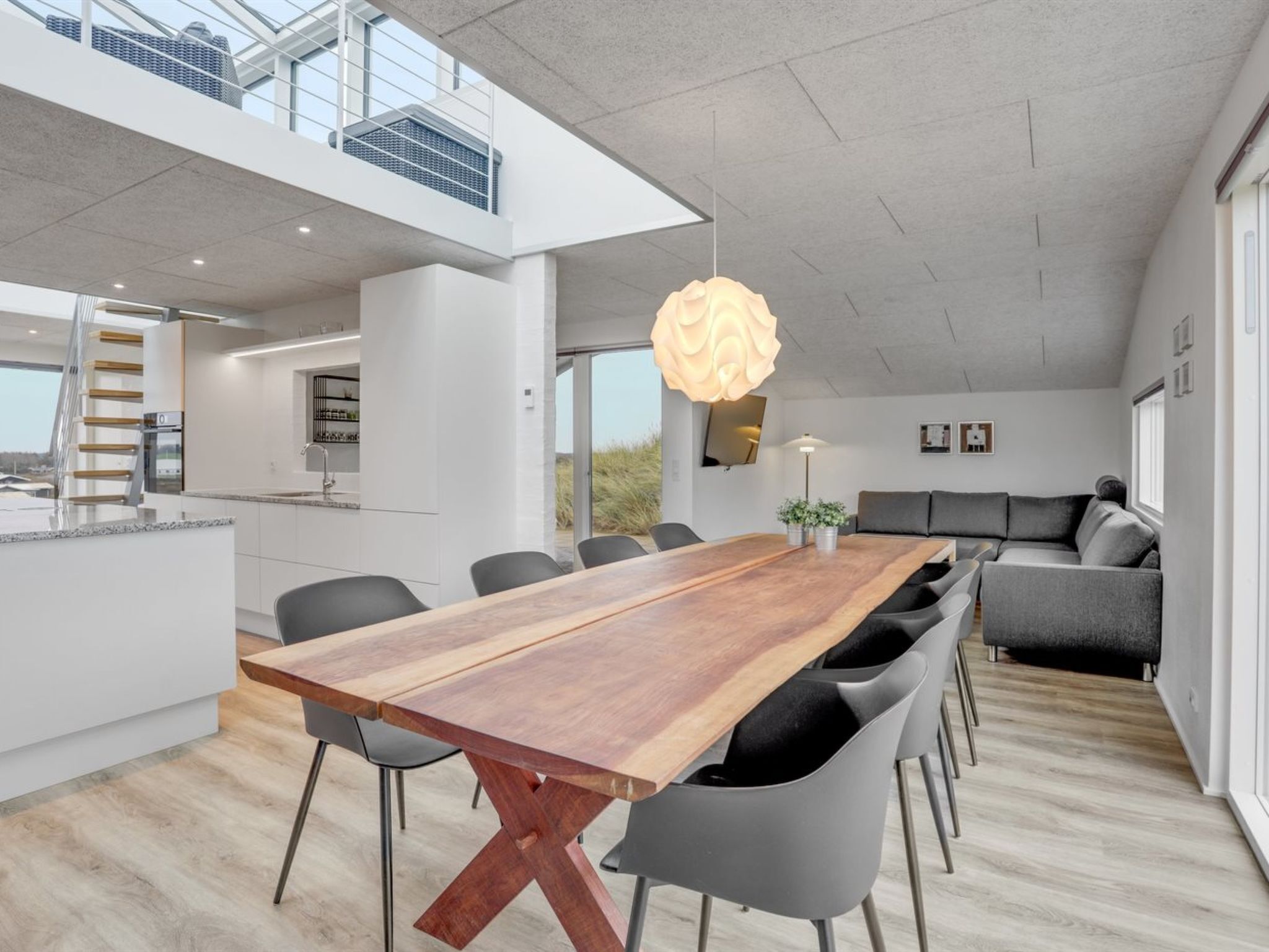 A modern interior with a long wooden dining table, grey chairs, a grey sofa, and a large window.