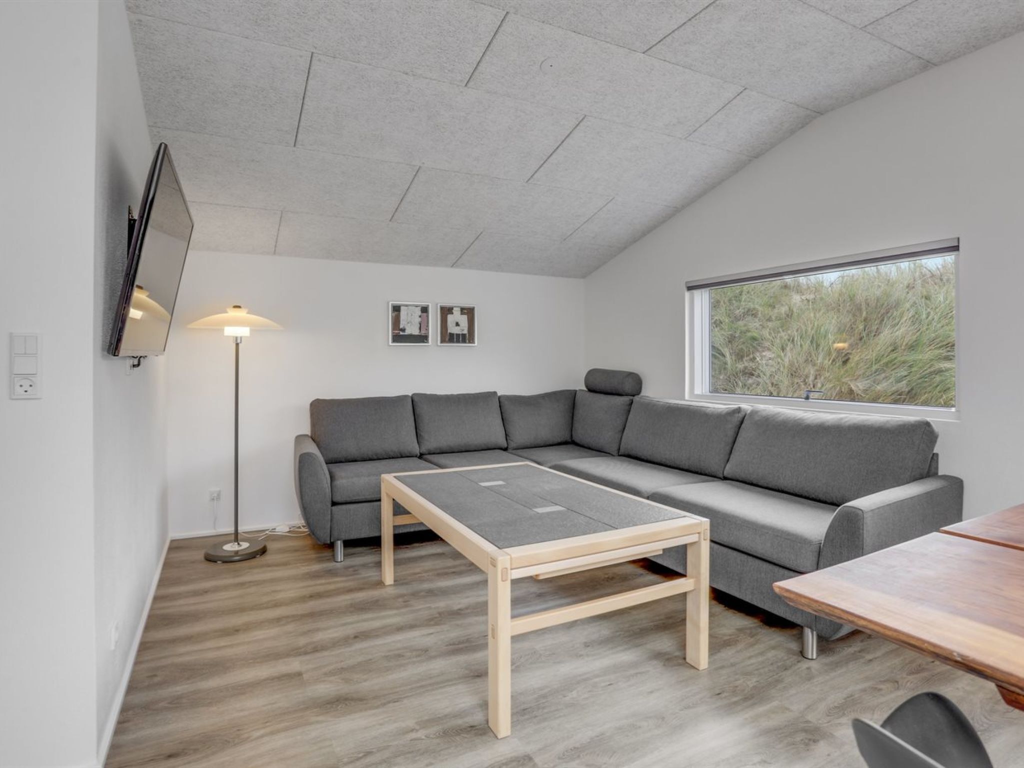 Ein modernes Wohnzimmer mit einem grauen Ecksofa, einem Holztisch, einer Stehlampe und einem an der Wand befestigten Fernseher.