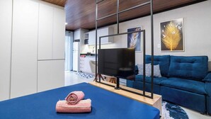 Ein modernes Wohnzimmer mit einem blauen Sofa, einem Flachbildfernseher auf einem Schrank, einer Holzzahlende und einer Küche im Hintergrund.