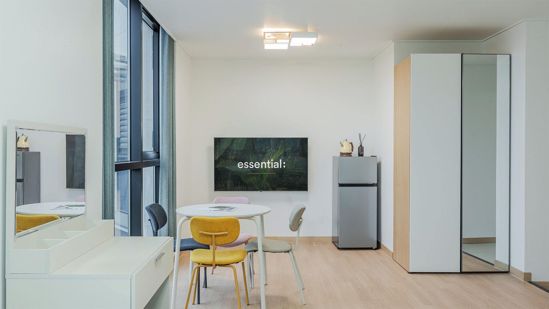 A modern, minimalist room with a large mirror, a small round table with chairs, a grey wardrobe, and a wall-mounted TV displaying the word 'essential'.