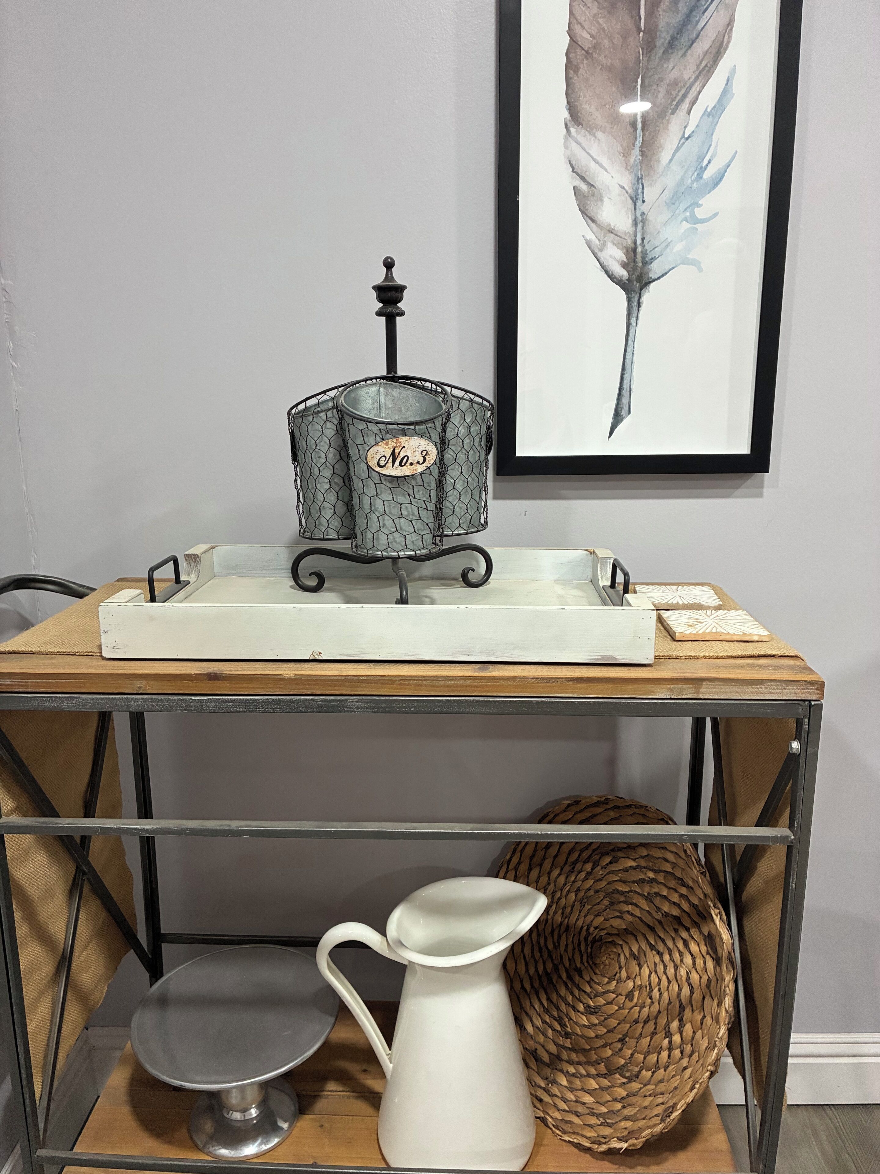 Una mesa con estructura metálica, una bandeja blanca, un jarro metálico y una cesta tejida sobre una superficie de madera.