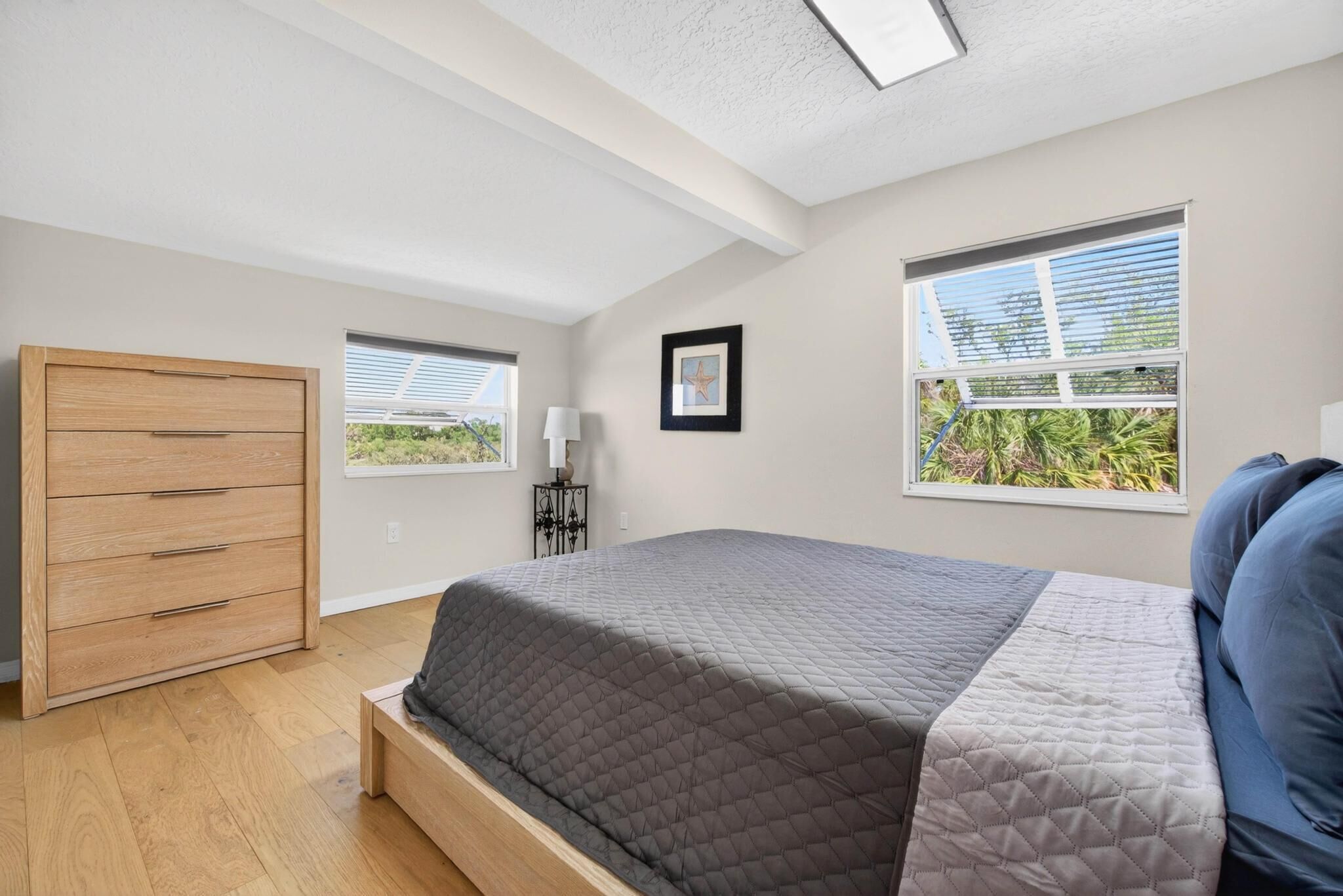 Une chambre à coucher avec un lit, une commode en bois, deux fenêtres équipées de stores et un luminaire au plafond.
