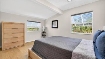 Une chambre à coucher avec un lit, une commode en bois, deux fenêtres équipées de stores et un luminaire au plafond.