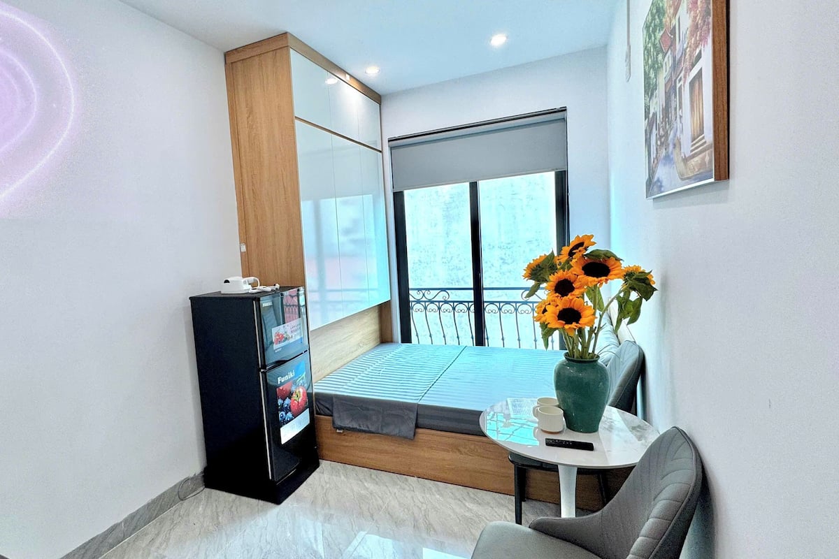 A modern room with a sofa, a small table with a vase of sunflowers, a black cabinet with a Funitel logo, and a balcony with a view.
