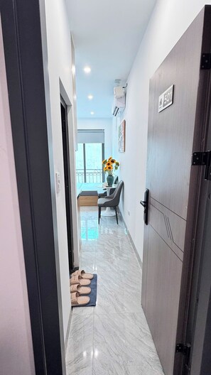 Um corredor com uma porta identificada como '501', uma pequena mesa com um vaso de girassóis e vista para uma varanda.