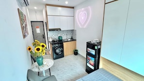A compact living space with a kitchen, a dining area with a table and chairs, a washing machine, and a refrigerator.