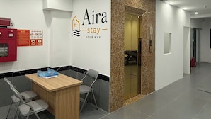Uma área de recepção de hotel, com balcão, duas cadeiras, uma placa fixada na parede e uma porta que dá para outro espaço.
