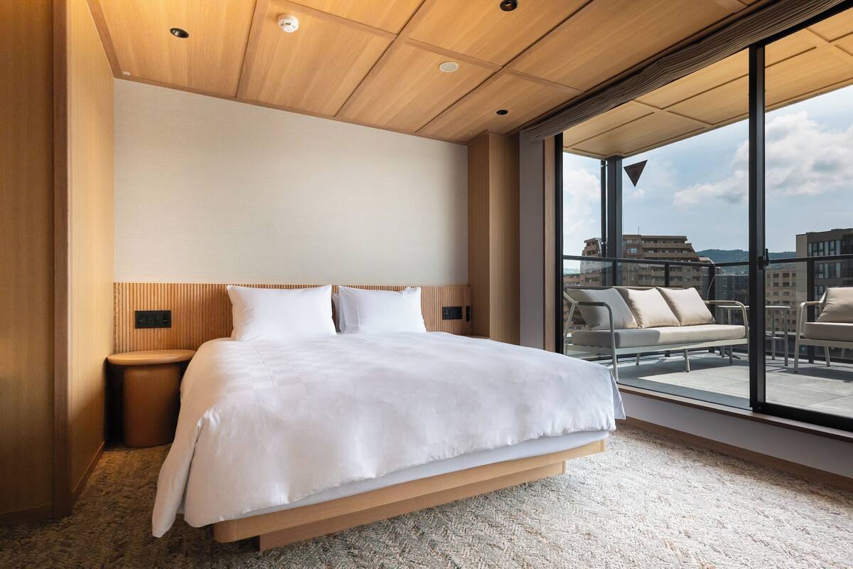 Una habitación de hotel moderna con una cama grande, una mesita de noche pequeña, un sofá y un balcón con vista a la ciudad.