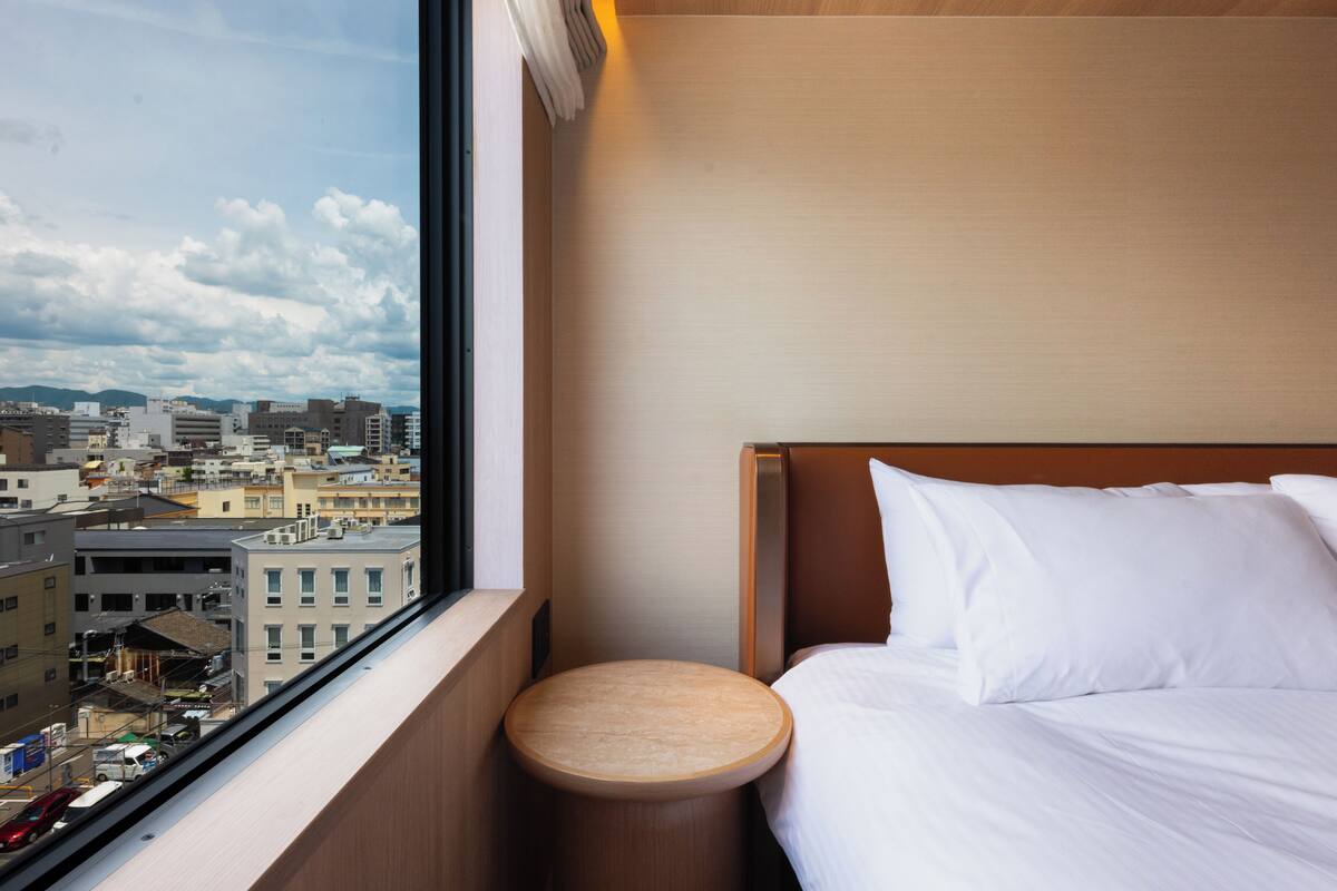 Habitación de hotel con cama, un taburete de madera redondo, una ventana con vista a la ciudad y cabecero de madera.