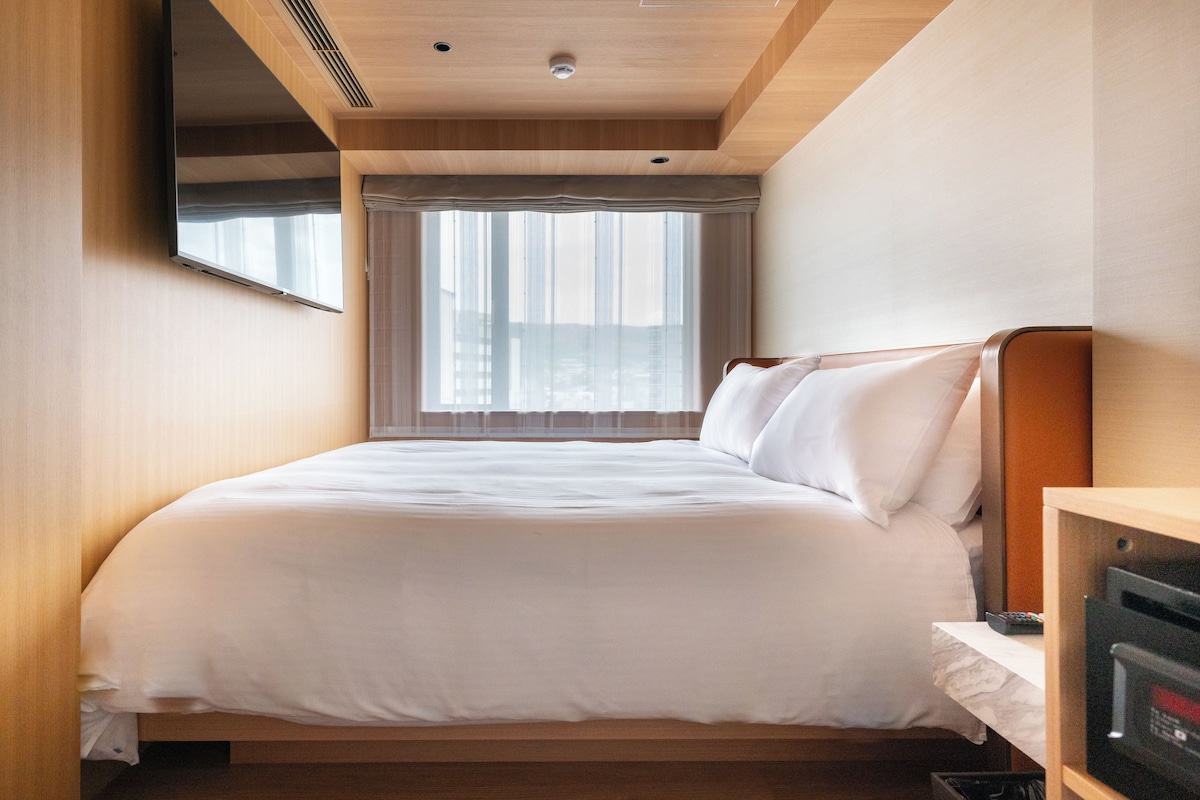 A hotel room with a bed, a television, a window with curtains, and a wooden ceiling.