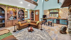 A cozy living room with a sectional sofa, a coffee table, and a fireplace. There are bookshelves and a kitchen area visible.
