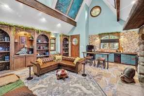 A cozy living room with a sectional sofa, a coffee table, and a fireplace. There are bookshelves and a kitchen area visible. - Romantic Storybook Cottage Hot Tub No Cleaning Fee (Lonoke)