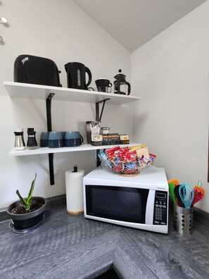 A kitchen with a white microwave, shelves with black kitchenware, a potted plant, and a basket of snacks.