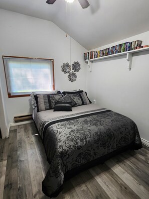 A bedroom with a bed, a ceiling fan, a window with blinds, a wall-mounted shelf with books, and a decorative wall piece.