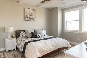 Quarto com cama, mesa de cabeceira, abajur, relógio e uma grande janela.