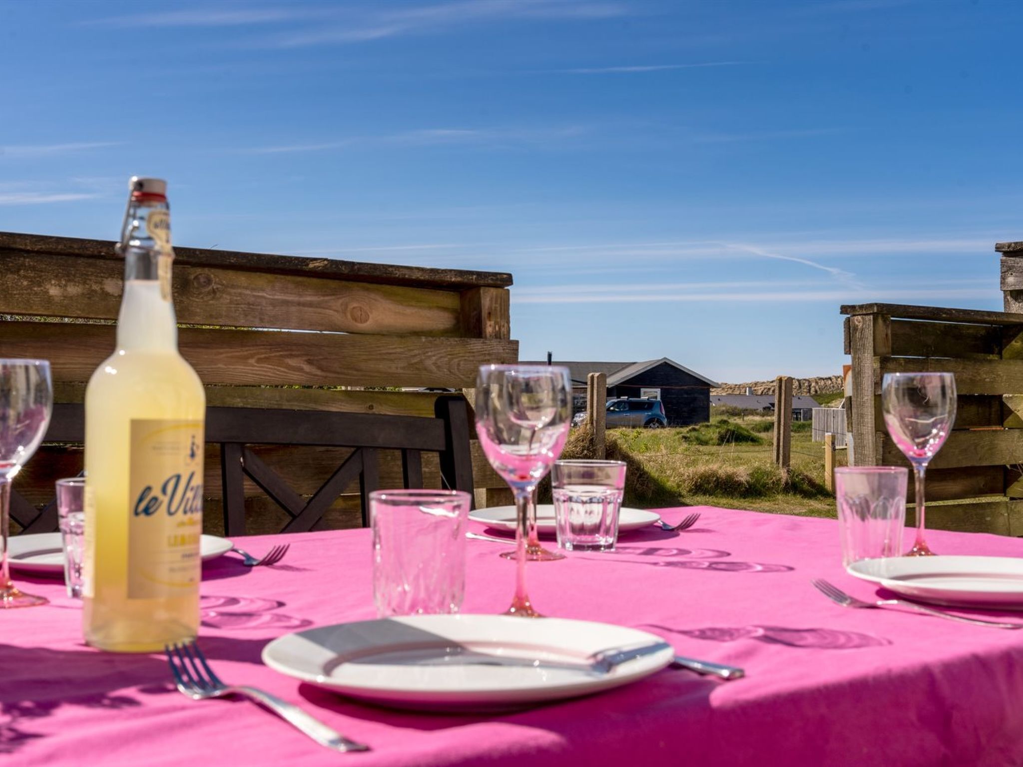 Une table dressée avec des verres à vin, une bouteille et des assiettes, une clôture en bois et un bâtiment en arrière-plan.