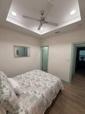 Ein Schlafzimmer mit einem Bett, Deckenventilator, Einbauleuchten, einem Spiegel und einer Tür.