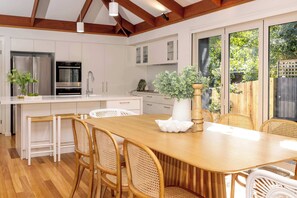 A modern kitchen with white cabinetry, stainless steel appliances, and a wooden dining table with wicker chairs. - The Mudjimba Beach House - 150m to the beach (Coolum Region)