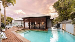 Ein moderner Poolbereich im Freien mit Holzterrasse, Liegestühlen und einer überdachten Sitzgelegenheit.