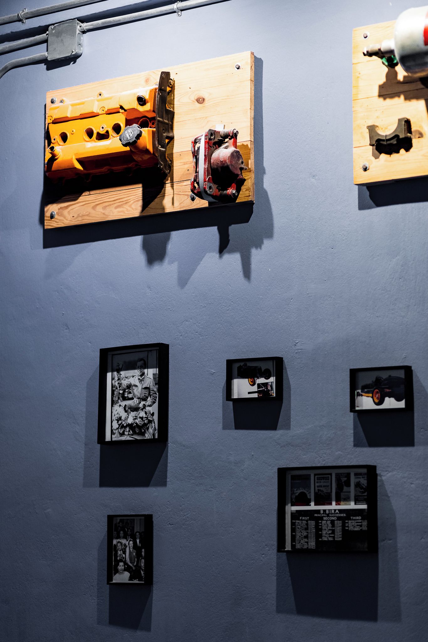 Una pared con varios objetos enmarcados, incluyendo un panel de madera con piezas mecánicas y varias fotografías en blanco y negro.