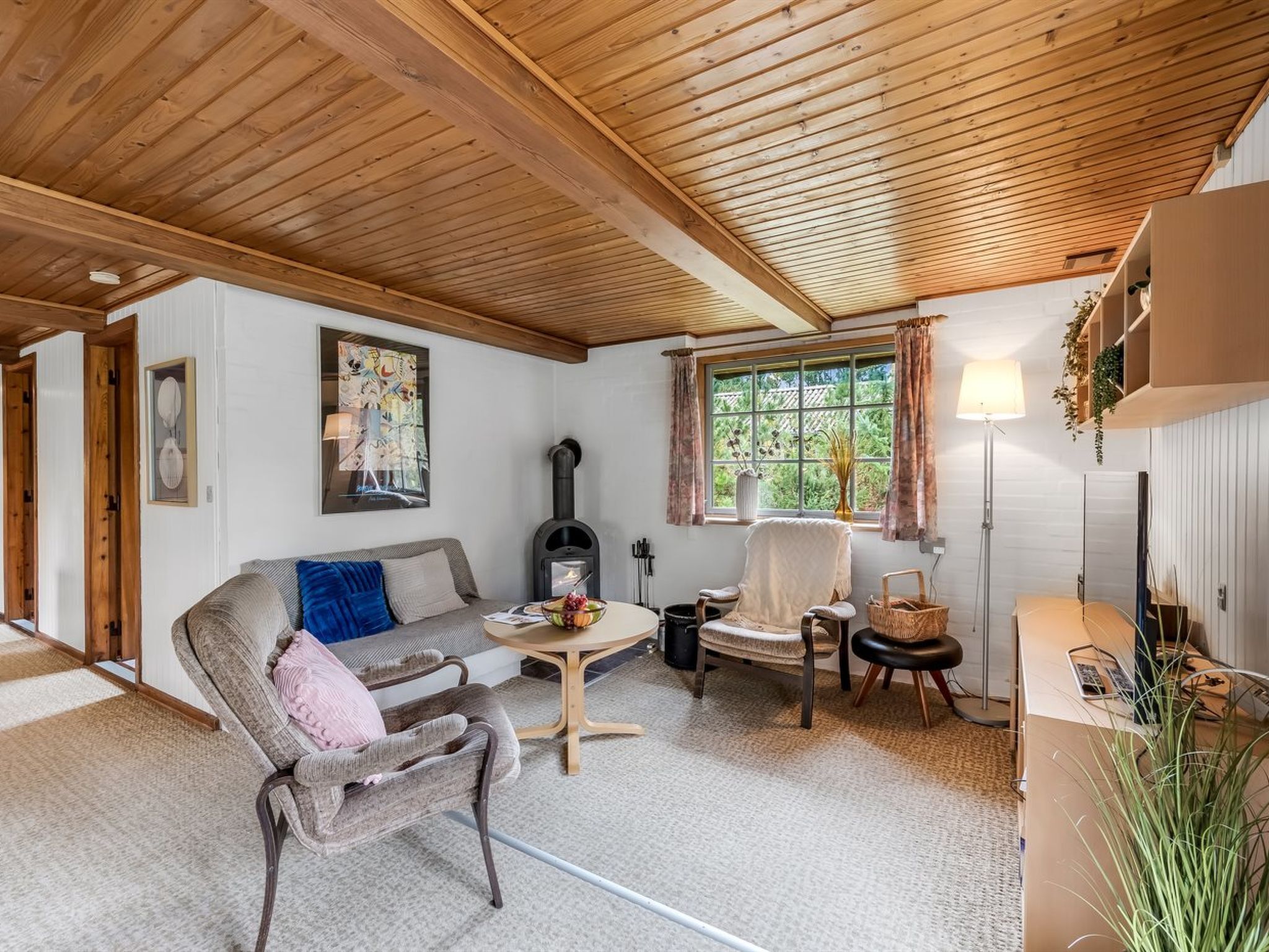Un salón acogedor con sofá, sillón y una mesa con un cuenco de fruta. Hay una chimenea, una ventana con cortinas y una lámpara.