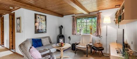Un salón acogedor con sofá, sillón y una mesa con un cuenco de fruta. Hay una chimenea, una ventana con cortinas y una lámpara.