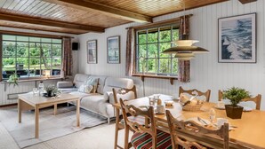 Une pièce chaleureuse avec un plafond en bois, un canapé, une table à manger avec des chaises et de grandes fenêtres avec des rideaux.